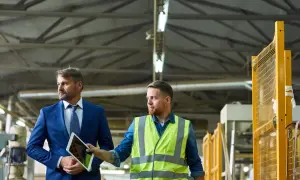 Dwóch mężczyzn w fabryce, jeden w garniturze z tabletem, drugi w kamizelce BHP.