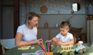 Mama i syn grają w gry planszowe. Dziecko układa drewniane pionki, a mama trzyma jednego w dłoni. Na stole kolorowe kredki i książeczka do kolorowania. Www.co to jest?