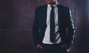 Mężczyzna w eleganckim garniturze z rękami w kieszeniach na tle ciemnej ściany.