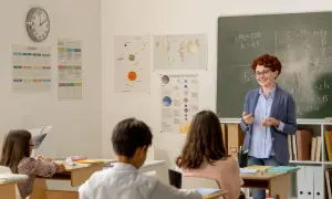 Nauczycielka z rudymi włosami tłumaczy coś uczniom przy tablicy w klasie.