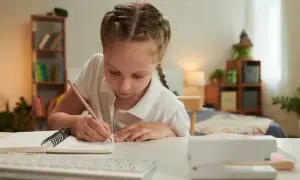 Dziewczynka z warkoczem pisze w zeszycie. Zastanawia się, czy za szkołę w chmurze się płaci, gdy odrabia lekcje w domu.