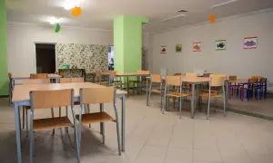Stołówka szkolna z drewnianymi stołami i krzesłami, ozdobiona kolorowymi balonami i obrazkami owoców.