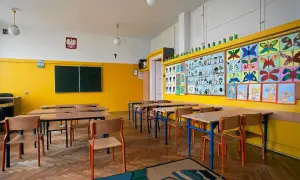Pusta klasa szkolna z żółtymi ścianami, ławkami, krzesłami i kolorowymi pracami dzieci.