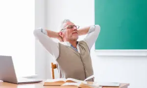 Nauczyciel zrelaksowany przy biurku, z rękami za głową, obok laptopa i książek.
