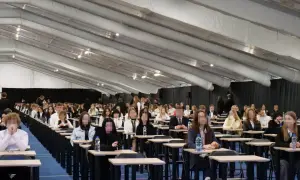Uczniowie siedzą w rzędach przy ławkach w dużej sali podczas egzaminu.