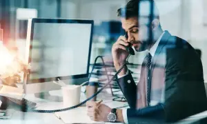 Biznesmen w garniturze rozmawia przez telefon i notuje w zeszycie przy biurku.