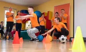 Dzieci i nauczyciele w pomarańczowych koszulkach ćwiczą z piłkami i pachołkami.