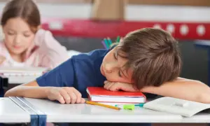 Chłopiec śpi na książce przy biurku w szkole, obok leży jabłko i ołówek.