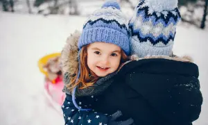 Uśmiechnięta dziewczynka w zimowej czapce przytulona do kogoś w tle, na śniegu.