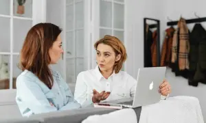 Dwie kobiety rozmawiają przy laptopie w jasnym pomieszczeniu.