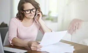 Kobieta w okularach rozmawia przez telefon, przeglądając dokumenty. Prawdopodobnie zajmuje się sprostowaniem świadectwa pracy.