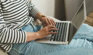 Kobieta w pasiastym swetrze i jeansach pracuje na laptopie, co symbolizuje nienormowany czas pracy i elastyczność.