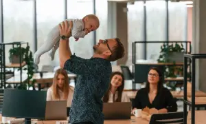 Mężczyzna w okularach podrzuca dziecko w biurze, gdzie pracują kobiety.