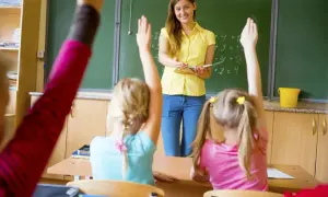 Nauczycielka w żółtej bluzce stoi przed tablicą, a uczniowie podnoszą ręce.