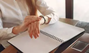 Kobieta sprawdza zegarek, zastanawiając się, ile płatne są nadgodziny. Na stole notatnik i laptop.