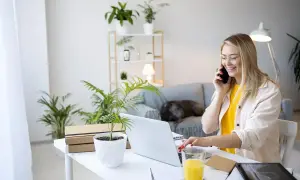 Uśmiechnięta kobieta rozmawia przez telefon przy biurku z laptopem i roślinami.