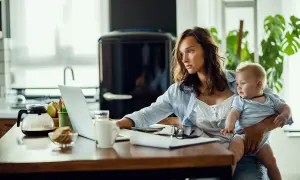 Kobieta z dzieckiem na rękach pracuje przy laptopie, przygotowując wniosek o urlop macierzyński.