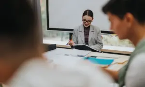 Kobieta w okularach i marynarce siedzi przy biurku, trzymając długopis i notatnik.