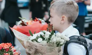 Chłopiec w białej koszuli trzyma bukiet białych kwiatów, na tle innych bukietów i ludzi.