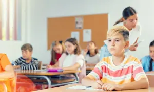 Uczniowie siedzą w klasie, a nauczycielka stoi z tyłu.