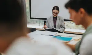 Nauczycielka w okularach i marynarce siedzi przy biurku z dokumentami, trzymając długopis.