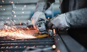 Pracownik w rękawicach szlifuje metal, sypią się iskry. Uważaj na wypadek przy pracy!