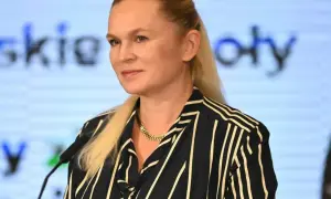 Kobieta z blond włosami w pasiastym ubraniu stoi przed mikrofonem na tle ekranu z tekstem.