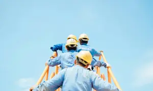 Czterech robotników w żółtych kaskach i niebieskich kombinezonach wspina się po pomarańczowych schodach. Stosują środki ochrony zbiorowej, dbając o bezpieczeństwo.