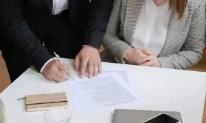 Dwie osoby przy stole podpisują dokumenty. Jedna z nich pisze piórem.