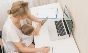Kobieta z dzieckiem na kolanach sprawdza na telefonie, urlop macierzyński ile płatny. Obok laptop i notatnik.