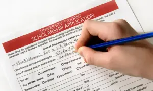 Dłoń pisze niebieskim długopisem na formularzu wniosku o stypendium uniwersyteckie.