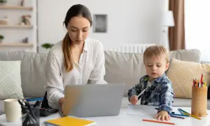 Kobieta pracuje przy laptopie, podczas gdy jej syn rysuje obok. To obrazek z życia, gdzie praca i rodzina łączą się, jak w przypadku art. 188 kodeksu pracy.