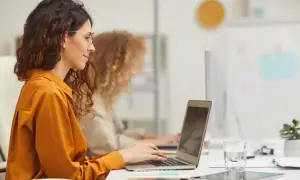 Kobieta w okularach, jako zleceniobiorca, pracuje przy laptopie w biurze. Obok niej siedzi koleżanka.