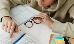 Osoba w bluzie z kapturem przegląda książkę, obok leżą okulary i kolorowe karteczki.