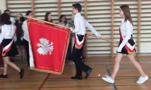 Uczniowie w galowych strojach tworzą poczet sztandarowy, niosąc biało-czerwony sztandar z godłem Polski.