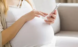 Ciężarna kobieta w białej bluzce i beżowym kardiganie używa telefonu komórkowego.