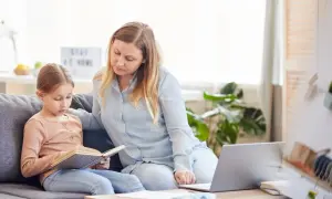 Kobieta i dziecko siedzą na kanapie, dziecko czyta książkę, obok leży laptop.