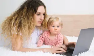 Kobieta z dzieckiem na rękach pracuje przy laptopie, zastanawiając się, czy na urlopie wychowawczym można pracować.