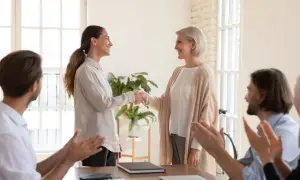 Uśmiechnięte kobiety ściskają sobie dłonie, celebrując jubileusz 30 lat pracy. Koledzy klaszczą.