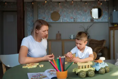Mama i syn grają w gry planszowe. Dziecko układa drewniane pionki, a mama trzyma jednego w dłoni. Na stole kolorowe kredki i książeczka do kolorowania. Www.co to jest?