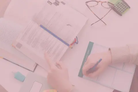 Otwarta książka, ręka pisząca w notatniku, okulary i kalkulator.