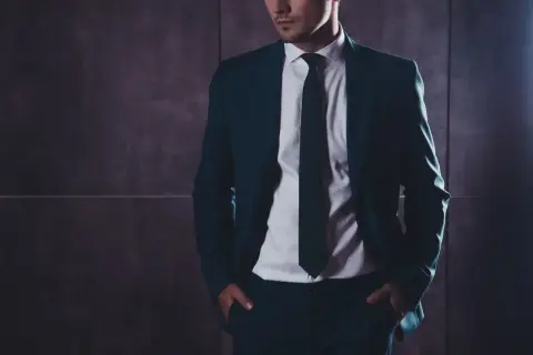 Mężczyzna w eleganckim garniturze z rękami w kieszeniach na tle ciemnej ściany.