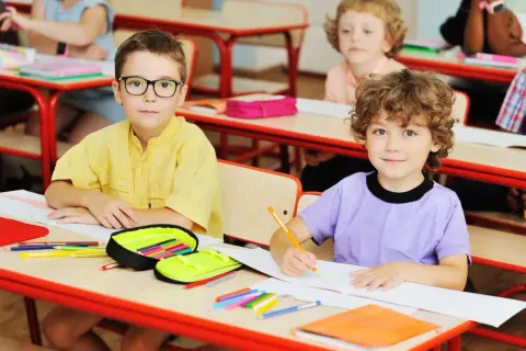 Dzieci w szkole siedzą przy ławkach, rysują i piszą.