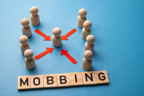 Drewniane figurki ludzi otaczają jedną, a czerwone strzałki wskazują na nią. Pod spodem litery układają się w słowo "MOBBING".