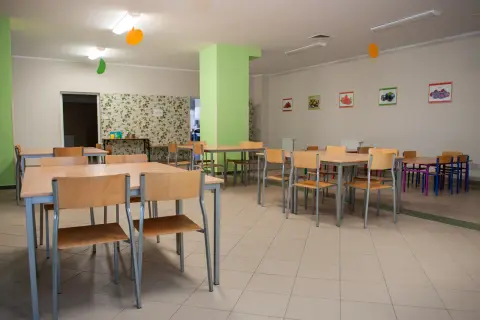Stołówka szkolna z drewnianymi stołami i krzesłami, ozdobiona kolorowymi balonami i obrazkami owoców.