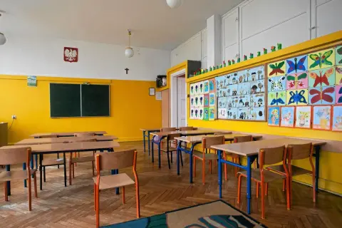 Pusta klasa szkolna z żółtymi ścianami, ławkami, krzesłami i kolorowymi pracami dzieci.