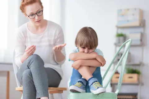 Kobieta w okularach rozmawia ze smutnym chłopcem siedzącym na krześle.
