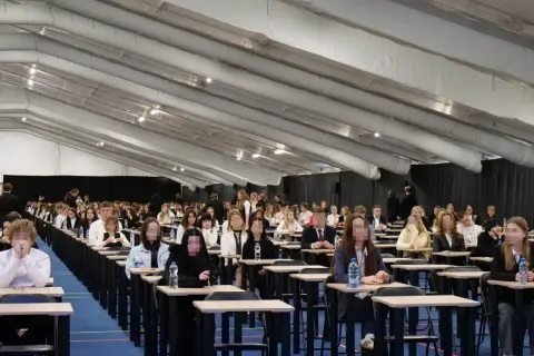 Uczniowie siedzą w rzędach przy ławkach w dużej sali podczas egzaminu.