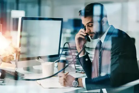 Biznesmen w garniturze rozmawia przez telefon i notuje w zeszycie przy biurku.