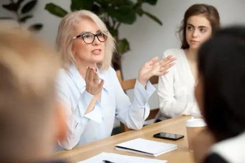 Kobieta w okularach gestykuluje podczas spotkania z innymi osobami przy drewnianym stole.